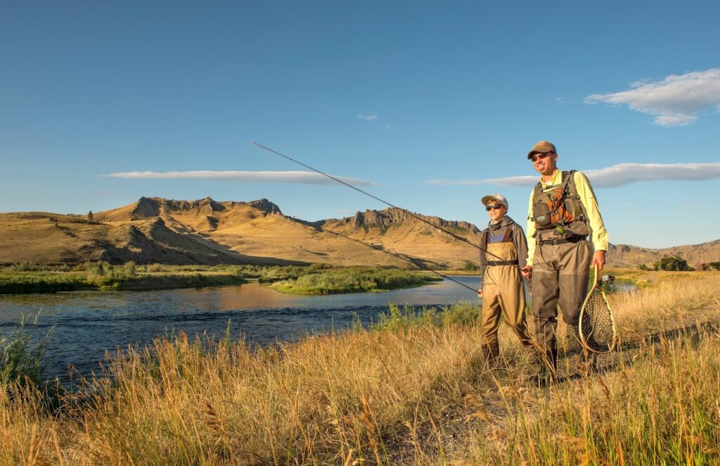 A father and son carry fishing poles through a grassy area next to a river, with mountains in the background in Great Falls, MT