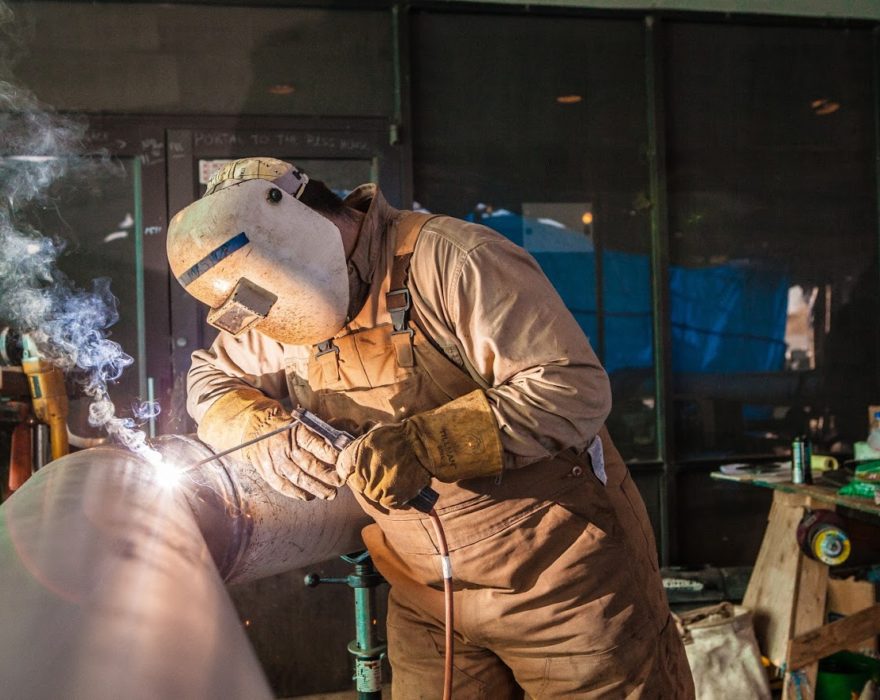 A welder with a face shield and gloves works on welding a piece of metal equipment