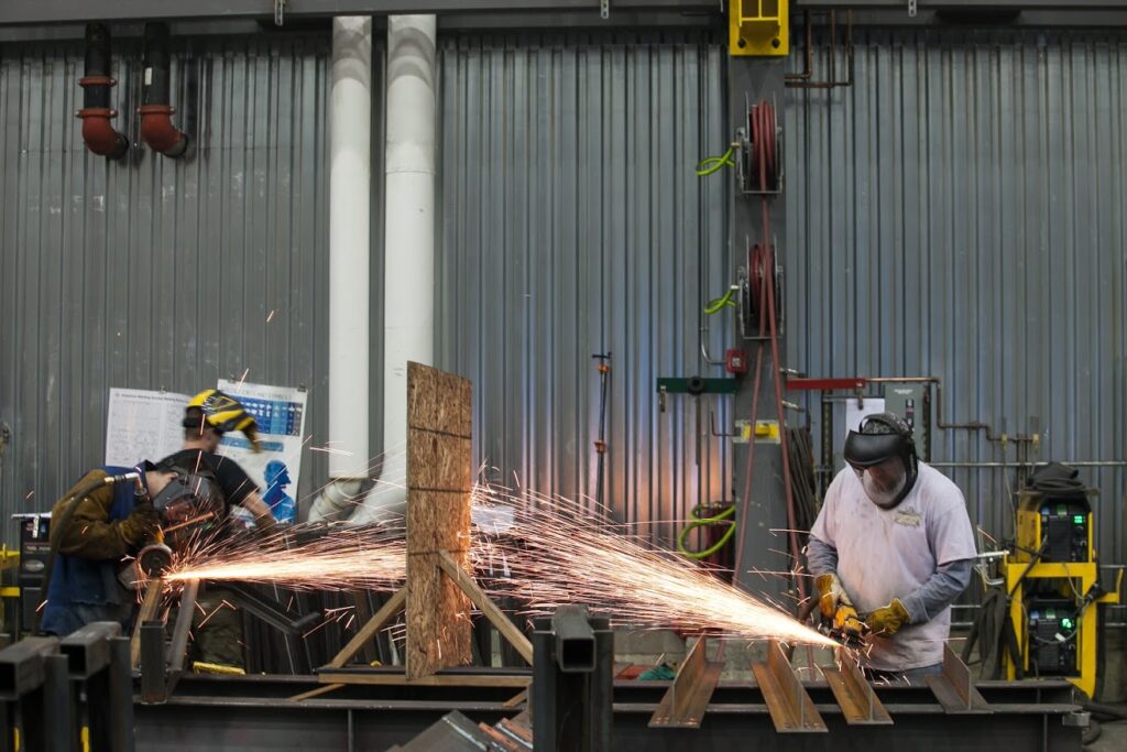Two men are welding in a warehouse with face shields and equipment
