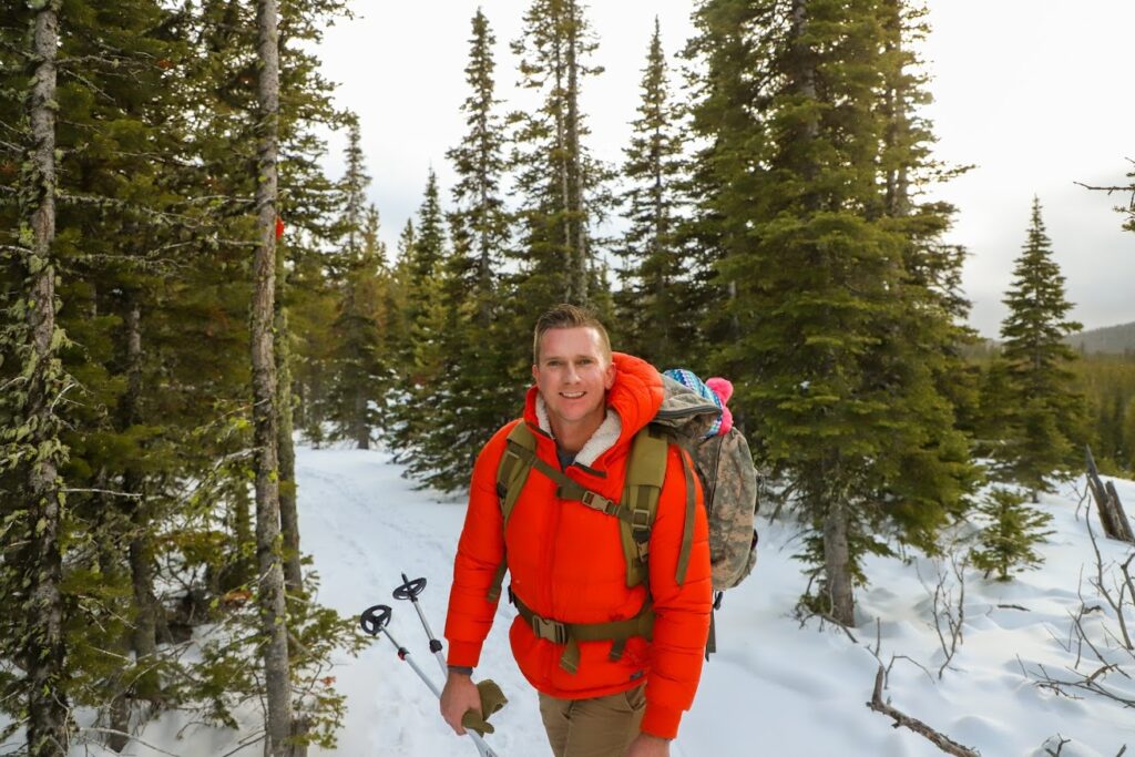 A man wearing an orange winter coat stops to smile for a photo in front of a background of snow and pine trees in Great Falls, MT