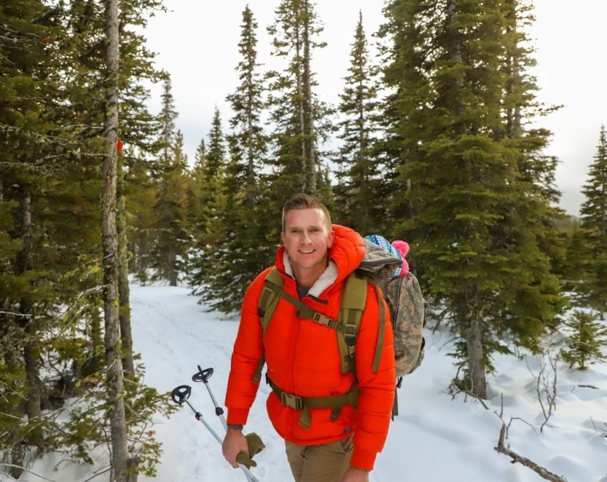 A man wearing an orange winter coat stops to smile for a photo in front of a background of snow and pine trees in Great Falls, MT