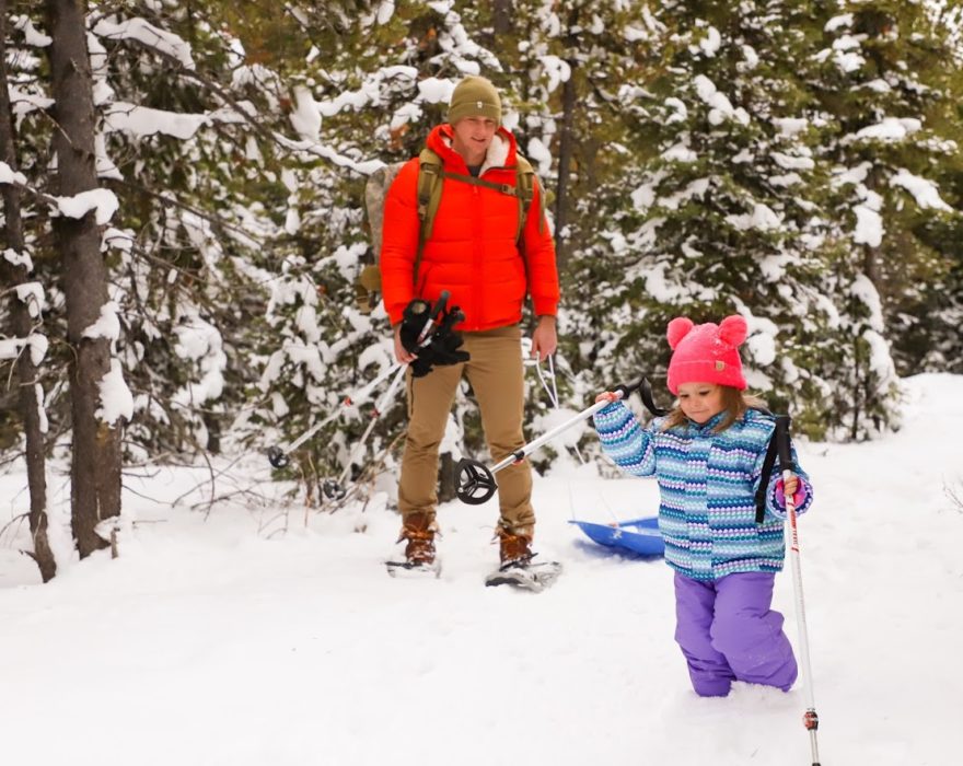 A child and her father use ski poles for support as they walk through the snow, with the father carrying a sled behind them